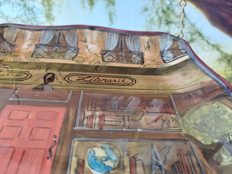 May include: A hand-painted ceramic wall hanging depicting a French bookstore with the word "Librairie" above the door. The bookstore has a red door, bookshelves, and a globe. The hanging is decorated with a gold chain.