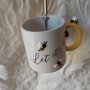 May include: White ceramic mug with a yellow handle and bee decorations. The mug has the text "Let it..." printed on the front.