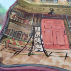 May include: A ceramic plate with a painted scene of a bookstore. The plate features a red door with six panels, a bookshelf filled with books, and the word "Library" written on the building.