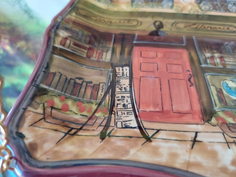 May include: A ceramic plate with a painted scene of a bookstore. The plate features a red door with six panels, a bookshelf filled with books, and the word "Library" written on the building.