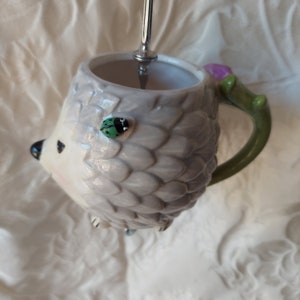 May include: A white ceramic mug shaped like a hedgehog with a green handle and a green ladybug on the side.