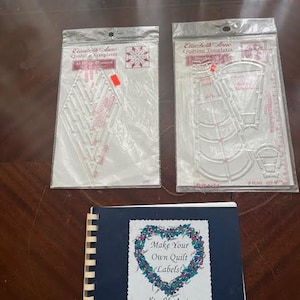 May include: A collection of quilting supplies, including clear plastic templates and a spiral-bound notebook. The notebook cover features a heart-shaped floral design with the text "Make Your Own Quilt Labels!"