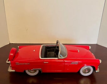May include: A red 1950s Ford Thunderbird convertible toy car model. The car has a white interior and white wheels.