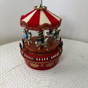 May include: A miniature carousel music box featuring a red and white striped canopy, a central rotating pole, and several painted horses. The base is circular, colored in deep red with gold accents, and surrounded by a small white railing.