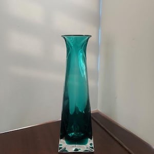May include: A teal blue glass vase with a square base and a narrow, tall opening.