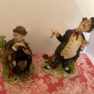 May include: Two ceramic figurines. One depicts a seated man playing a violin, wearing a brown hat and coat. The other shows a man in a suit gesturing with a dog at his feet. Both are on decorative bases with gold accents.