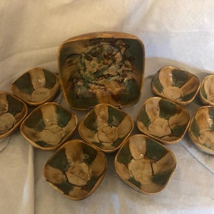 May include: A set of nine ceramic bowls in shades of brown, green, and white. The bowls are square and have a rustic, handmade look. The set includes a larger bowl and eight smaller bowls.