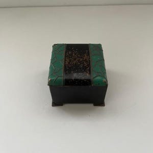 May include: A small, rectangular black box with a hinged lid. The lid is decorated with green textured panels on the sides and a central black panel with a speckled, brown design. The box has a simple, minimalist design.
