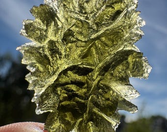 May include: A translucent, olive-green gemstone with a rough, textured surface. The natural stone is held between fingers against a backdrop of a blue sky. The gemstone has a unique, organic shape with multiple facets, catching the light.