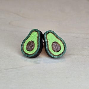 May include: A pair of avocado-shaped stud earrings. Each earring features a green outer layer, a brown seed, and a dark green border. The earrings are made of wood and are designed to be worn as jewelry.