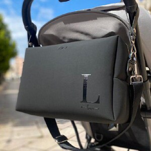 Könnte beinhalten: Eine schwarze Wickeltasche mit einem großen Buchstaben L auf der Vorderseite, befestigt an einem schwarzen Kinderwagen.