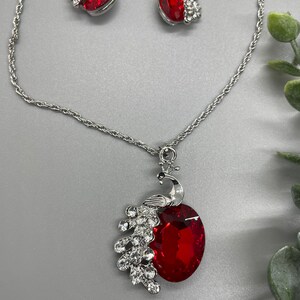 May include: A silver necklace with a red crystal peacock pendant and matching earrings. The pendant features a detailed peacock design with clear crystal accents.