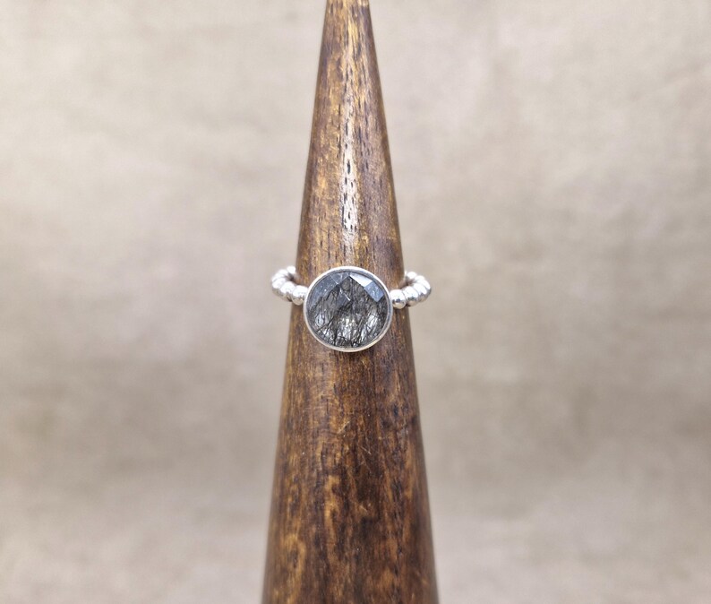 May include: A silver ring with a large, oval-shaped, black and gray stone. The band has a textured, beaded design. The ring is displayed on a wooden cone-shaped stand against a neutral background.
