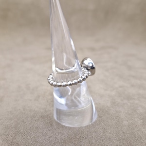 May include: A silver ring with a beaded band and a smooth, rounded top. The ring is displayed on a clear, conical stand against a neutral background. The ring's design is simple and elegant.