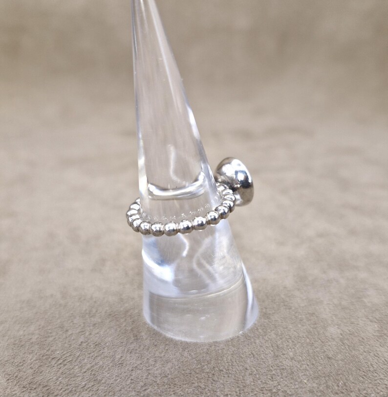 May include: A silver ring with a beaded band and a smooth, rounded top. The ring is displayed on a clear, conical stand against a neutral background. The ring's design is simple and elegant.