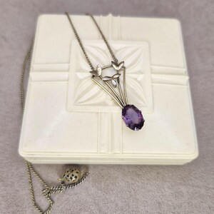May include: A silver-toned necklace with a pendant featuring a large, oval-cut purple gemstone. The pendant has a stylized floral design with three leaves. The necklace rests on a white, textured box.