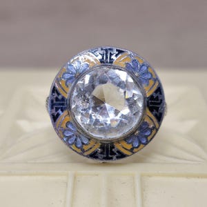 May include: A circular ring featuring a large, clear, faceted centre stone surrounded by a decorative border of blue floral patterns and yellow accents set against a dark, geometric background within a metallic frame.