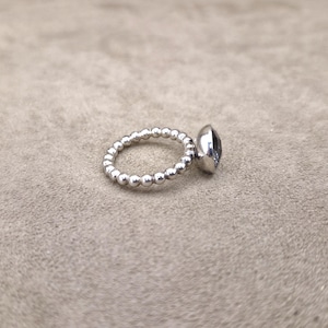 May include: A silver ring with a beaded band and a large, oval-shaped, dark-colored gemstone. The ring is set against a neutral background, showcasing its polished metal and the gem's facets.