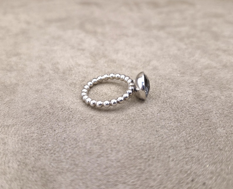 May include: A silver ring with a beaded band and a large, oval-shaped, dark-colored gemstone. The ring is set against a neutral background, showcasing its polished metal and the gem's facets.
