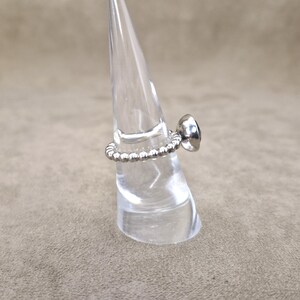 May include: A silver ring with a beaded band and a dark, possibly black, gemstone. The ring is displayed on a clear, cone-shaped stand against a neutral background. The gemstone is round and set in a smooth bezel.