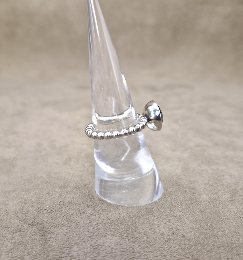 May include: A silver ring with a beaded band and a dark, possibly black, gemstone. The ring is displayed on a clear, cone-shaped stand against a neutral background. The gemstone is round and set in a smooth bezel.