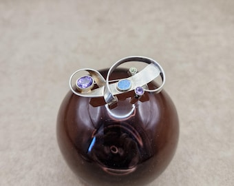 May include: A silver brooch with an abstract design featuring a swirl of metal and several small gemstones in shades of purple, blue, and green.