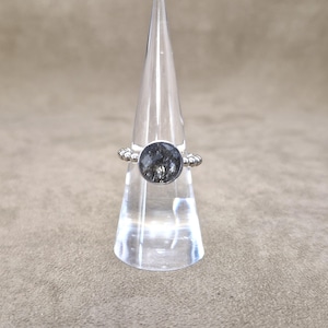 May include: A silver ring with a large, round, dark gray stone. The ring is displayed on a clear, cone-shaped stand. The band of the ring has a decorative, beaded design. The stone has a unique, textured appearance.