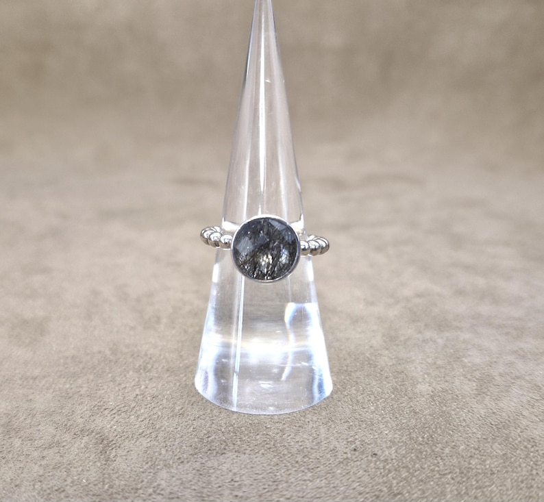May include: A silver ring with a large, round, dark gray stone. The ring is displayed on a clear, cone-shaped stand. The band of the ring has a decorative, beaded design. The stone has a unique, textured appearance.