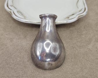 May include: A small, teardrop-shaped silver-toned metal object with a smooth, reflective surface and a narrow neck opening at the top. The item has a rounded, bulbous base and a polished finish that catches light, creating bright highlights on its curved exterior.