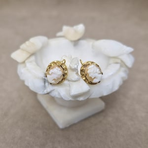 May include: A pair of gold-coloured cameo earrings. Each earring features a carved profile within an oval frame with a floral design. The cameos have a light peach hue, set against a gold background. The earrings are displayed on a white, sculpted decorative piece.