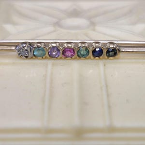 May include: A silver-toned bar brooch featuring a horizontal row of seven small, round, multi-colored gemstones. The stones display a gradient of colors including light blue, teal, purple, pink, green, dark blue, and dark grey, set in a metal frame.