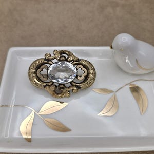 May include: An antique-style brooch with a large, clear, oval-shaped crystal center set in an ornate, gold-toned metal frame. The brooch rests on a white rectangular dish with gold leaf accents, next to a small white ceramic bird.