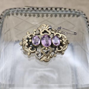 May include: An antique brooch with three oval, light purple gemstones set in an ornate, gold-toned metal frame. The brooch features a decorative, swirling design and a safety pin clasp.