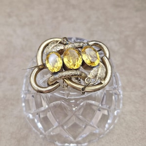 May include: A vintage brooch featuring three oval, yellow gemstones set in a gold-toned metal frame. The frame has an ornate, swirling design with leaf accents. The brooch is secured with a pin clasp.