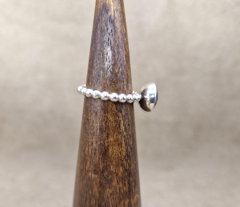 May include: A silver ring with a beaded band and a round, dark-colored stone. The ring is displayed on a wooden ring holder against a neutral background. The ring's design is simple and elegant.