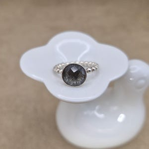 May include: A silver ring with a large, round, faceted, dark gray gemstone with black inclusions. The ring band is made of small silver beads. The ring is displayed on a white, flower-shaped ceramic dish.