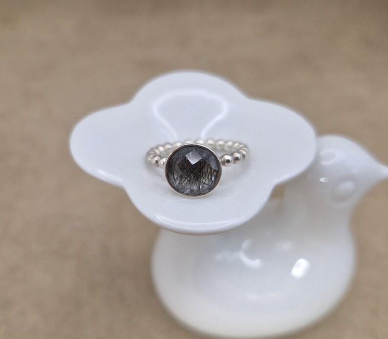 May include: A silver ring with a large, round, faceted, dark gray gemstone with black inclusions. The ring band is made of small silver beads. The ring is displayed on a white, flower-shaped ceramic dish.
