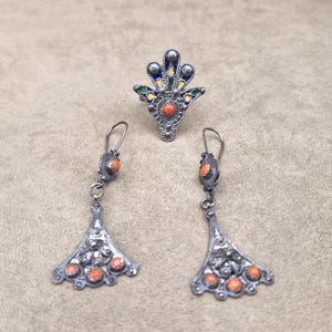 May include: A silver-toned jewelry set featuring a ring and a pair of earrings. The ring has a fan-shaped design with blue, green, and yellow enamel accents and a central orange stone. The earrings have a similar design with orange stones.