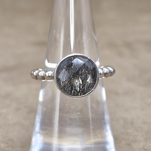 May include: A silver ring with a round, faceted, black and clear gemstone. The band features a beaded design. The ring is displayed on a clear, cone-shaped stand against a neutral background. Jewelry.