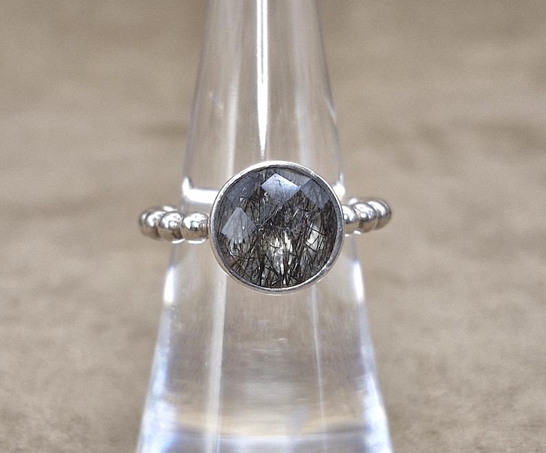 May include: A silver ring with a round, faceted, black and clear gemstone. The band features a beaded design. The ring is displayed on a clear, cone-shaped stand against a neutral background. Jewelry.