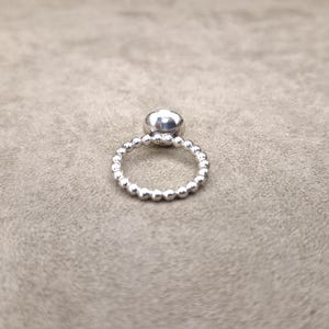 May include: A silver ring with a large, round, clear gemstone. The band is made of small, evenly spaced silver beads. The ring is set against a neutral, textured background.