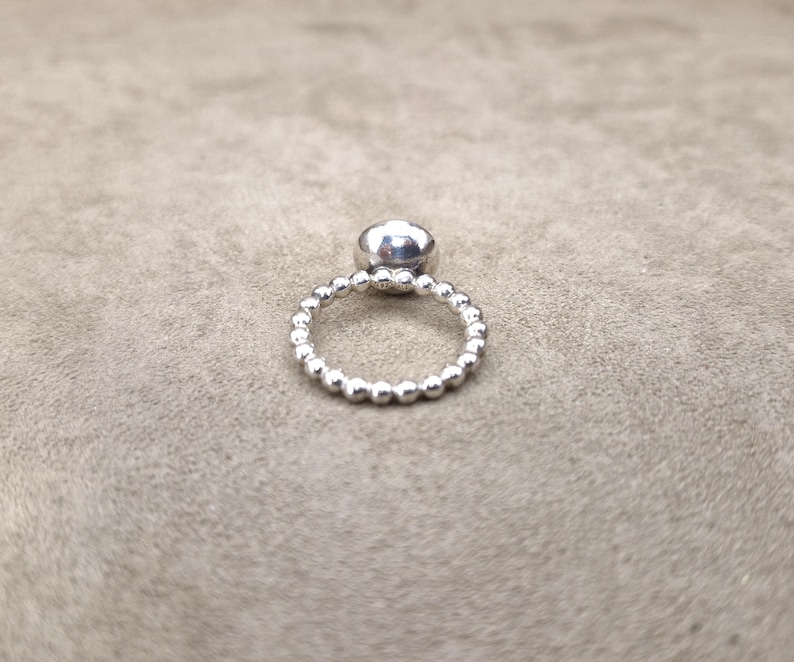 May include: A silver ring with a large, round, clear gemstone. The band is made of small, evenly spaced silver beads. The ring is set against a neutral, textured background.