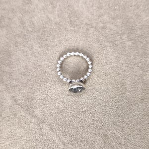 May include: A silver ring with a beaded band and a large, clear, faceted gemstone. The ring is set against a neutral, textured background, highlighting its simple elegance. The ring's design is classic and suitable for everyday wear.