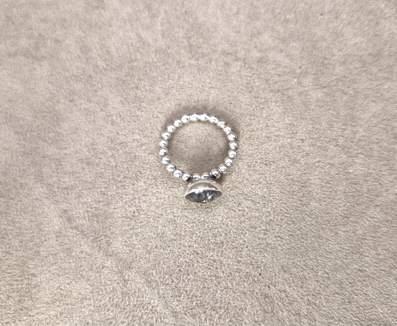 May include: A silver ring with a beaded band and a large, clear, faceted gemstone. The ring is set against a neutral, textured background, highlighting its simple elegance. The ring's design is classic and suitable for everyday wear.
