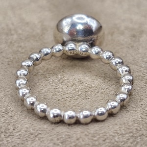 May include: A silver ring with a large, rounded top and a band of small, spherical beads. The ring is made of shiny, polished metal. The inscription "S925 ALE" is visible on the band. The ring is displayed on a neutral-toned surface.