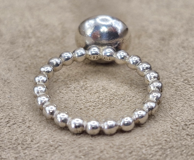 May include: A silver ring with a large, rounded top and a band of small, spherical beads. The ring is made of shiny, polished metal. The inscription "S925 ALE" is visible on the band. The ring is displayed on a neutral-toned surface.