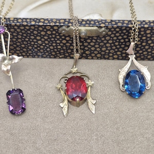 May include: Three metal necklaces featuring decorative pendants. The left pendant has a purple rectangular stone and a leaf motif. The center pendant features a large oval red stone with ornate metalwork. The right pendant holds an oval blue stone.
