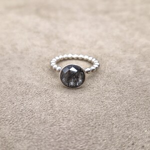 May include: A silver ring with a large, round, faceted black gemstone. The band is detailed with small, evenly spaced silver beads. The ring is set against a neutral, textured background.