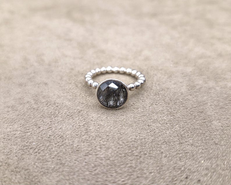 May include: A silver ring with a large, round, faceted black gemstone. The band is detailed with small, evenly spaced silver beads. The ring is set against a neutral, textured background.