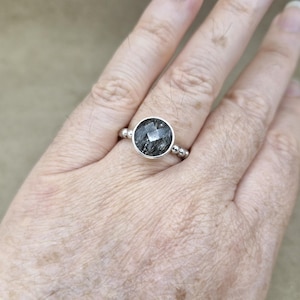 May include: A silver ring featuring a large, round, faceted black gemstone. The ring has a simple band with small silver beads on either side of the gemstone setting. The gemstone has a unique, textured appearance.